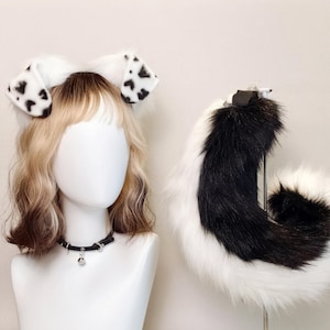 May include: A headband with white ears and black spots, and a fluffy tail. The tail is black and white, and the headband is on a mannequin head with a black collar and a silver bell.