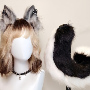 K&ouml;nnte beinhalten: Ein Set aus Kunstfell-Tierohren in Grau und Wei&szlig; mit silbernen Sternanh&auml;ngern, einem schwarzen Halsband mit einer kleinen silbernen Glocke und einem langen, geschwungenen schwarz-wei&szlig;en Kunstfellschwanz auf einem wei&szlig;en Schaufensterpuppenkopf.