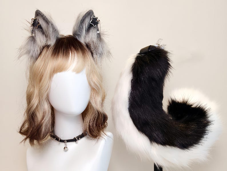 K&ouml;nnte beinhalten: Ein Set aus Kunstfell-Tierohren in Grau und Wei&szlig; mit silbernen Sternanh&auml;ngern, einem schwarzen Halsband mit einer kleinen silbernen Glocke und einem langen, geschwungenen schwarz-wei&szlig;en Kunstfellschwanz auf einem wei&szlig;en Schaufensterpuppenkopf.