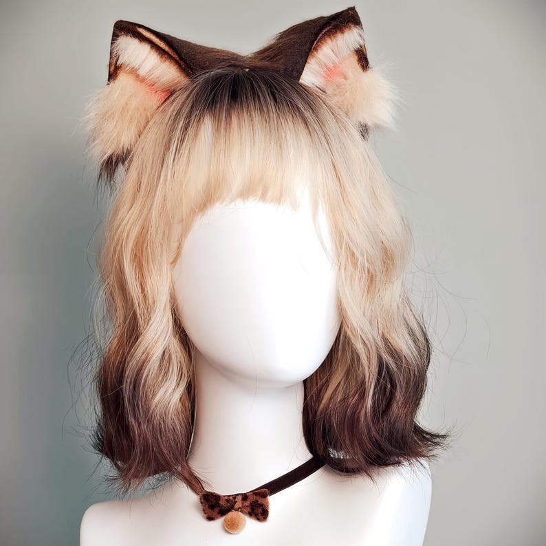 May include: A shoulder-length wig with blonde roots and dark brown ends, styled with wavy texture and bangs. The wig is adorned with brown and cream-colored faux fur animal ears. A thin brown velvet choker with a small bow and bell is included.