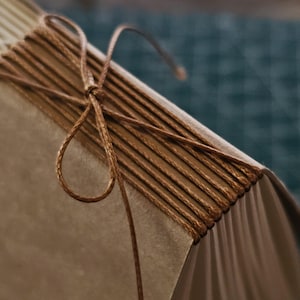 Könnte beinhalten: Nahaufnahme eines handgefertigten Journals mit braunem Einband und Seiten. Der Rücken ist mit braunem Faden handgenäht, und oben ist eine dekorative Schleife gebunden. Die Seiten des Journals sind sichtbar, was auf ein leeres Notizbuch oder Skizzenbuch hindeutet.