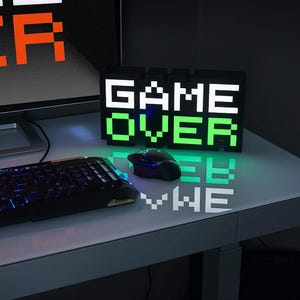 May include: A black LED light sign displaying "GAME OVER" in pixelated white and green text. A black computer mouse and keyboard are in the foreground. The sign is on a white desk.