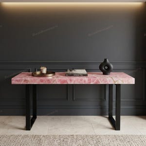 May include: A rectangular console table featuring a polished pink rose quartz stone top with natural white veining, supported by two black metal U-shaped legs. A small candle on a tray, two stacked books, and a black abstract vase sit on the surface.