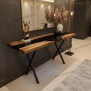 May include: A modern console table with a live edge wood top and black epoxy resin inlay. The table has a black metal base with an X-shaped design. A gold sculpture and decorative vase are on the table.