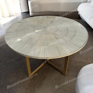 May include: A round coffee table featuring a white mother-of-pearl inlay top with a concentric circular pattern. The table is supported by a gold-coloured metal base with intersecting legs that form an X-shape beneath the circular tabletop surface.