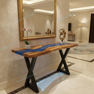 May include: A wooden console table with a live edge design and a blue resin river. The table has black metal X-shaped legs. The table is in a well-lit entryway with a mirror and decorative items.