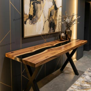 May include: A wooden console table with a black epoxy river design. The table has a natural wood grain finish and sits on a black metal X-shaped base. Two bronze vases and a painting hang in the background.