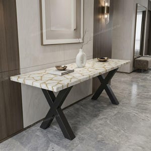 May include: A rectangular console table featuring a white surface with a gold-toned crackled pattern and black metal X-shaped legs. A white vase and two small metallic bowls are placed on the tabletop, along with a book with a white cover.