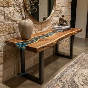 Può includere: Una consolle in legno con un fiume di resina blu e pietre al centro. Il tavolo ha un bordo naturale e poggia su gambe in metallo nero. Un vaso con rami e libri sono sopra.