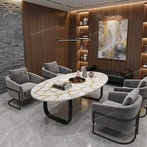 Op de afbeelding: Een ovale eettafel met een wit en goud marmerpatroon blad en een zwart metalen onderstel, omringd door vier grijze gestoffeerde fauteuils met zwarte metalen frames. Een stapel boeken en een glazen karaf staan op het tafeloppervlak.