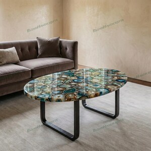 Puede incluir: Una mesa de centro ovalada con un tablero compuesto por numerosas rodajas de ágata pulidas y multicolores en tonos azul, verde, marrón y blanco. La mesa se apoya sobre una base de metal oscuro en forma de U con un acabado mate y liso.