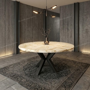 Op de afbeelding: Een ronde eettafel met een gepolijst crèmekleurig en beige marmeren blad met natuurlijke aders. De tafel wordt ondersteund door een zwart metalen onderstel met kruisende poten. Een kleine metalen vaas staat in het midden van het blad.