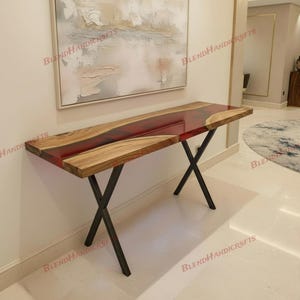 Puede incluir: Una consola artesanal con una tapa de madera de borde vivo y una incrustación de resina roja vibrante. La mesa está soportada por patas de metal negro con un diseño en forma de X. La mesa mide aproximadamente 152 cm de largo.