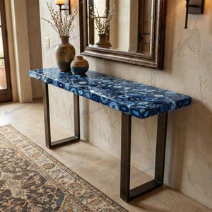 Puede incluir: Mesa consola rectangular con tapa de ágata azul y patas de metal oscuro. La encimera presenta un mosaico de rodajas de ágata azul y blanca. En el fondo se ven jarrones decorativos y un espejo.