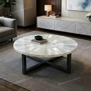 May include: A round coffee table featuring a white, iridescent, sunburst-patterned tabletop made of inlaid material. The table is supported by a dark, matte-finished metal base with a cross-legged design that provides structural stability.