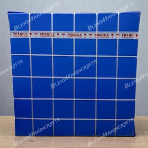 May include: A blue box with a grid pattern and white lines. The box is sealed with tape that reads "FRAGILE HANDLE WITH CARE" with a graphic of a wine glass. The box has the words "BLEND HANDICRSFTS" printed on it.