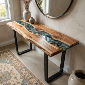 May include: A wooden console table with a river-like design. The table features a live edge wooden top with a blue epoxy resin river filled with stones. The table is supported by black metal legs. The table measures approximately 60 inches long.