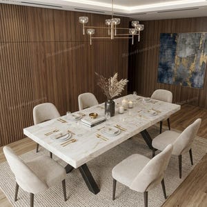 May include: A rectangular dining table with a white marble top and black metal legs, surrounded by six beige upholstered chairs. The table is set with white plates, gold cutlery, glassware, and a central vase with dried foliage.