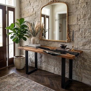 May include: A wooden console table with a black epoxy river design. The table has black metal legs and is placed against a stone wall. A gold-framed mirror hangs above the table, reflecting the room. A potted plant and vase with dried flowers are also visible.