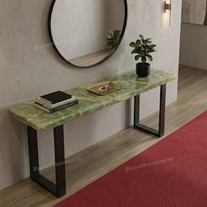 May include: A rectangular console table featuring a polished green onyx stone top with natural veining and black metal U-shaped legs. The table surface holds a small stack of books and a gold-toned rectangular tray, with a potted plant nearby.