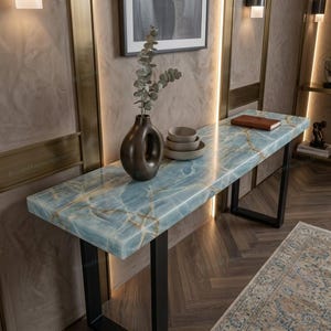 May include: A console table featuring a polished light blue stone top with gold veining and a thick rectangular profile. The table is supported by black metal legs and holds a dark brown ceramic vase with a branch, stacked bowls, and a notebook.