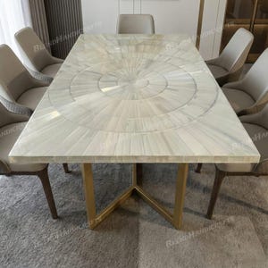 Könnte beinhalten: Ein rechteckiger Esstisch mit einer Oberfläche aus weißem Perlmutt-Inlay in einem konzentrischen Kreismuster. Der Tisch wird von einem goldfarbenen Metallgestell mit schrägen Beinen getragen und ist von sechs gepolsterten Stühlen in Hellbeige umgeben.