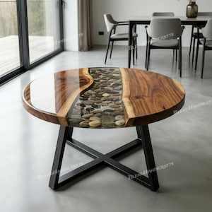May include: A round wooden coffee table with a unique design. The table features a clear resin river filled with small stones, set between two pieces of natural wood. The table has a black metal base.