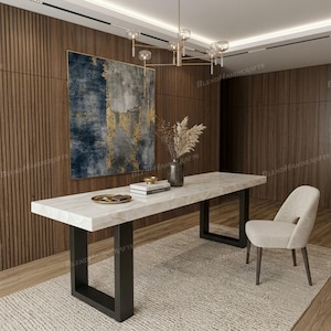 May include: A rectangular dining table with a white marble top and black U-shaped metal legs. A dark gray vase with dried foliage sits on the table next to a stack of books and a small gold tray. A single cream-colored upholstered chair is nearby.