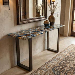 May include: A rectangular console table featuring a tabletop composed of multicolored blue, brown, and white agate slices with a polished finish. The table is supported by two dark metal U-shaped legs. Two ceramic vases sit on the table surface.