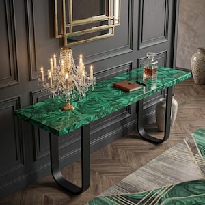 Puede incluir: Una mesa consola rectangular con una superficie de patrón de malaquita verde y dos patas de metal negro en forma de U. La mesa sostiene un candelabro de cristal con velas encendidas, un cuaderno de cuero marrón y un decantador de vidrio.