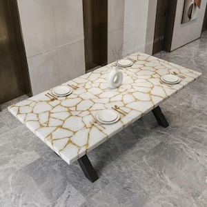 May include: A rectangular dining table featuring a white stone surface with a gold-toned crackled pattern, supported by black metal legs. The table is set with four white plates, gold-toned cutlery, and a white ceramic vase placed in the centre.