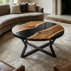 May include: Round coffee table with a black epoxy resin center and natural wood edges. The table has a dark metal X-shaped base. The wood grain is visible, and the table is set in a living room.