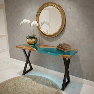 Op de afbeelding: Een houten console met een turquoise epoxyhars middenstuk en live edge details. De tafel heeft zwarte metalen poten en is versierd met een goudkleurige spiegel, een orchidee in pot en een stapel boeken. De tafel is ongeveer 122 cm lang.