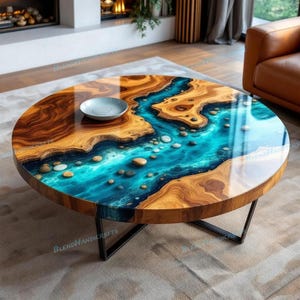 May include: Round wooden coffee table with a river-like design. The table features a clear epoxy resin river with blue and turquoise hues, embedded with small stones. The table has a dark wood border and sits on a black metal base.