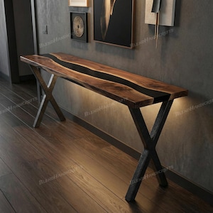 May include: A long console table featuring a natural live-edge wood slab surface with a central black resin river inlay. The table is supported by two black metal X-shaped legs and includes integrated under-table lighting that casts a soft glow.
