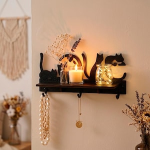 Könnte beinhalten: Dunkelbraunes Holzregal mit Katzen-Silhouette-Design. Es enthält eine brennende Kerze, ein Glas mit Lichterketten und kleine Vasen mit getrockneten Blumen. Darunter befinden sich Haken mit Schmuck.