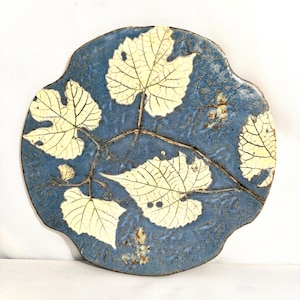 May include: A round, ceramic plate with a blue background and a leaf design. The leaves are light yellow with detailed veining, and the plate has a unique, scalloped edge. The plate is a decorative item.
