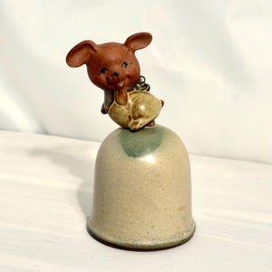 Może przedstawiać: Zabytkowy ceramiczny dzwonek z figurką brązowego misia na beżowej podstawie w kształcie dzwonka z zielonym akcentem. Miś ma duże uszy i trzyma mały przedmiot. Dzwonek to przedmiot dekoracyjny.