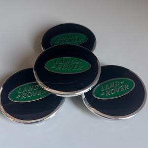 May include: Four black and silver Land Rover centre caps with a green logo. The caps are circular with a shiny finish and are arranged on a white surface. The Land Rover logo is in a green oval.