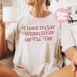Puede incluir: Una camiseta beige de manga corta con el texto granate I HAVE TO SAY WEIRD STUFF OR I'LL DIE impreso en una fuente serif estilizada en el pecho. La camiseta tiene un cuello redondo y un corte relajado, mostrada en un entorno casual.