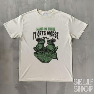 Könnte beinhalten: Cremefarbenes T-Shirt mit einem grünen Aufdruck von zwei Fröschen mit Cowboyhüten, die auf einem Blatt sitzen. Der Text "HANG IN THERE IT GETS WORSE" ist grün über den Fröschen aufgedruckt. Das Shirt hat einen Rundhalsausschnitt und kurze Ärmel.
