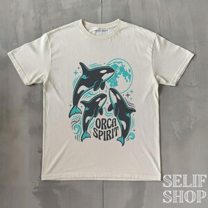 Könnte beinhalten: Cremefarbenes T-Shirt mit einer Grafik von drei Orcas mit türkisfarbenen Akzenten, einem türkisfarbenen Mond und den Worten "ORCA SPIRIT". Das Design beinhaltet Wellen und Sterne. Das Shirt hat kurze Ärmel und einen Rundhalsausschnitt.