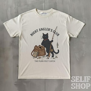 Op de afbeelding: Een crèmekleurig T-shirt met korte mouwen en een afbeelding van een zwarte kat die een hengel vasthoudt. De tekst luidt NIGHT ANGLER'S CLUB en THE PURR-FECT CATCH. De kat staat naast een bruine rieten vismand met kleine vissen en kunstaas.