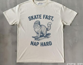May include: Cream-colored t-shirt featuring a blue illustration of a cat on a skateboard, wearing a beanie. The text "SKATE FAST, NAP HARD" is printed above and below the cat graphic. The shirt has short sleeves and a crew neck.