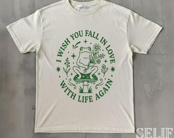Op de afbeelding: Crèmekleurig T-shirt met een groene afbeelding. De afbeelding toont een kikker met bloemen, zittend op een paddenstoel, met de tekst "I wish you fall in love with life again" eromheen.