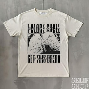 Könnte beinhalten: Cremefarbenes T-Shirt mit einer schwarz-weißen Grafik von zwei sich küssenden Tauben. Das Shirt hat japanischen Text über den Tauben und den Satz "GET THIS BREAD" darunter. Das Shirt hat einen Rundhalsausschnitt.