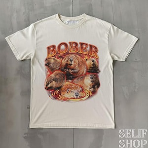 Könnte beinhalten: Cremefarbenes T-Shirt mit einer Grafik von mehreren Bibern und dem Wort "BOBER" in roter und orangefarbener Schrift. Die Biber sind kreisförmig angeordnet, mit einem wasserähnlichen Design um sie herum. Das Shirt hat kurze Ärmel.