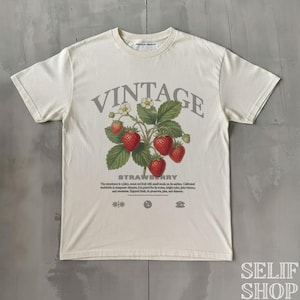 Può includere: T-shirt color crema con un design vintage a fragole. La grafica include la parola "VINTAGE" sopra un'illustrazione dettagliata di fragole, foglie e fiori. La parola "STRAWBERRY" è sotto, con testo aggiuntivo.