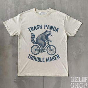 Könnte beinhalten: Cremefarbenes T-Shirt mit einer blauen Illustration eines Waschbären auf einem Fahrrad. Der Text "Trash Panda Trouble Maker" ist über und unter der Waschbären-Grafik aufgedruckt. Das Shirt hat kurze Ärmel und einen Rundhalsausschnitt.