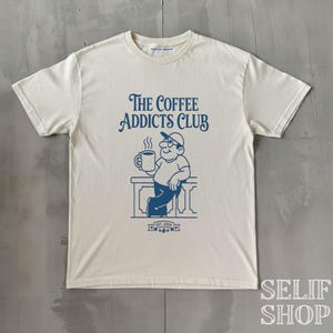 Op de afbeelding: Crèmekleurig T-shirt met een blauwe afbeelding van een man met een koffiemok, leunend op een balustrade. De tekst "The Coffee Addicts Club" staat boven de illustratie, met "EST. 2024" eronder. Het shirt heeft een ronde hals en korte mouwen.